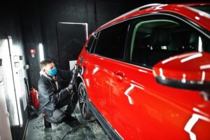 Technician wearing a mask cleans the wheel area of a red vehicle inside a detailing bay.