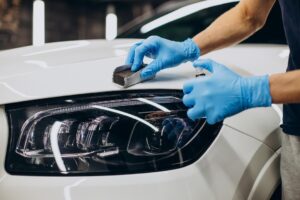 Detailer wearing blue gloves applies product near the headlight of a white vehicle during exterior detailing.