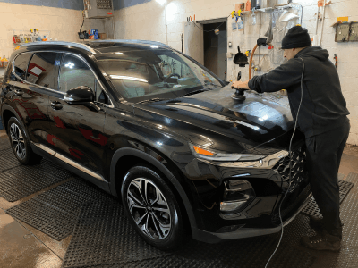 Black SUV before paint enhancement, showing a detailer polishing the hood inside a detailing garage.